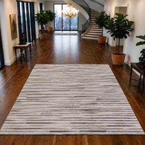 Op de afbeelding: Een rechthoekig vloerkleed met een horizontaal streeppatroon in verschillende tinten grijs, wit en antraciet. Het kleed ligt op een donkerbruine hardhouten vloer in een grote kamer met kamerhoge ramen en potplanten.