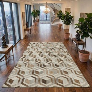 Op de afbeelding: Een rechthoekig vloerkleed met een geometrisch patroon van in elkaar grijpende zeshoeken en kubussen in crème, beige en bruine tinten. Het kleed ligt op een gepolijste houten vloer in een ruime hal met grote ramen en kamerplanten.