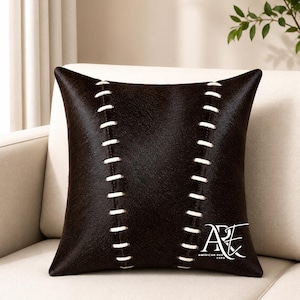 May include: A square dark brown throw pillow featuring a textured surface and two vertical rows of white whipstitching that resemble baseball seams. The bottom right corner displays a white logo with the text ARE AMERICAN RUG EXPO.