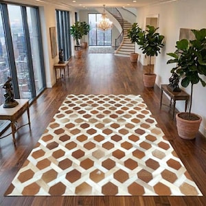Könnte beinhalten: Ein rechteckiger Teppich mit einem sich wiederholenden geometrischen Muster aus braunen und cremefarbenen sechseckigen Formen. Der Teppich liegt auf einem dunklen Holzboden in einem geräumigen Flur mit großen Fenstern, Pflanzen und einer Treppe.
