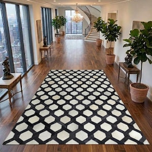 Könnte beinhalten: Ein rechteckiger Teppich mit einem sich wiederholenden schwarz-weißen hexagonalen geometrischen Muster. Der Teppich liegt auf einem dunklen Holzboden in einem geräumigen Flur mit großen Fenstern, Topfpflanzen und einer Treppe im Hintergrund.