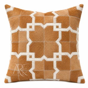 May include: A square throw pillow featuring a patchwork design of tan and brown hair-on hide leather. The surface is decorated with a geometric pattern formed by white leather strips stitched into the hide. A logo is visible in the bottom left corner.