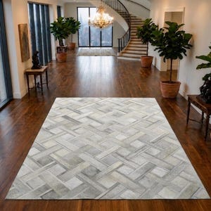 May include: A rectangular area rug featuring a woven-look pattern composed of light grey, white, and silver-toned panels. The rug displays a geometric grid design with overlapping rectangular segments that create a textured, multi-tonal appearance.