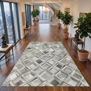 Puede incluir: Una alfombra rectangular con un patrón geométrico de cuadrados entrelazados en tonos gris, blanco y plateado. La alfombra está colocada sobre un suelo de madera oscura en un pasillo con grandes ventanales, plantas en macetas y una escalera.