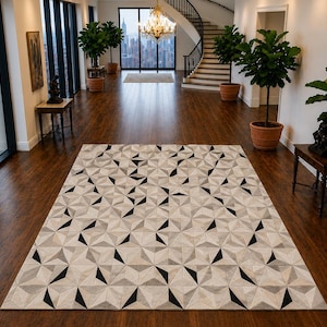 Op de afbeelding: Een rechthoekig vloerkleed met een herhalend geometrisch patroon van driehoeken in tinten crème, lichtgrijs en effen zwart. Het kleed ligt plat op een donkere houten vloer en toont een gestructureerd, meerkleurig ontwerp over het oppervlak.