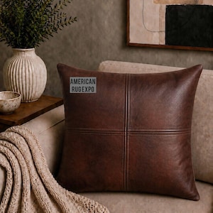 Peut inclure: Un coussin carré en cuir marron avec des détails de croix cousus. Le coussin est posé sur un canapé beige, avec une couverture en tricot beige et un vase de verdure en arrière-plan. Le texte "AMERICAN RUGEXPO" est visible sur le coussin.