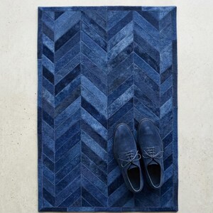 May include: A rectangular rug featuring a dark blue herringbone pattern made of stitched hide panels. A pair of matching dark blue suede lace-up shoes are placed on the right side of the rug, showcasing a consistent monochromatic color palette.