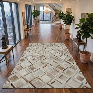 Op de afbeelding: Een rechthoekig vloerkleed met een geometrisch patroon van in elkaar grijpende vierkanten in crème, beige en bruine tinten. Het kleed ligt op een donkere houten vloer in een hal met grote ramen, potplanten en een trap.