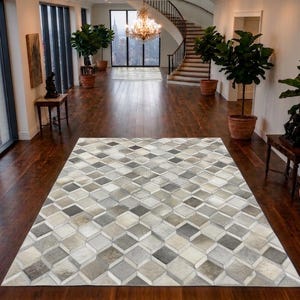 Puede incluir: Una alfombra rectangular con un patrón geométrico de formas de diamante entrelazadas en tonos gris, blanco y beige. La alfombra está compuesta por baldosas cuadradas individuales dispuestas para crear un efecto de cubo tridimensional.