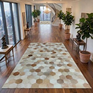 Op de afbeelding: Een rechthoekig vloerkleed met een herhalend geometrisch patroon van zeshoeken in verschillende tinten bruin, beige, crème en wit. Het kleed ligt op een donkere houten vloer in een ruime hal met grote ramen en kamerplanten.