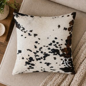 May include: A square throw pillow featuring a cowhide pattern with a white base and irregular black and brown spots. The lower right corner displays a white logo with the text ARE and American Rug Expo written in a stylized font.