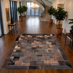 Puede incluir: Una alfombra rectangular con un diseño geométrico de patchwork compuesto por varios azulejos cuadrados y rectangulares en tonos gris carbón, gris pizarra, marrón y cobre metálico, dispuestos en un patrón repetitivo sobre un suelo de madera.