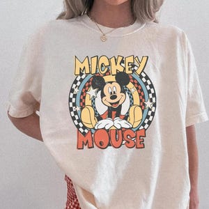 May include: Cream-colored t-shirt featuring a graphic of Mickey Mouse. The design includes the words "Mickey Mouse" in a retro font, set against a checkered and rainbow background. The shirt is paired with red and white checkered shorts.