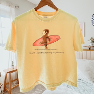 May include: A pale yellow short-sleeve t-shirt featuring a graphic of a brown monkey carrying a red surfboard. Below the graphic, the text reads, "I don't want this feeling to go away." The shirt is displayed on a wooden hanger against a light background.