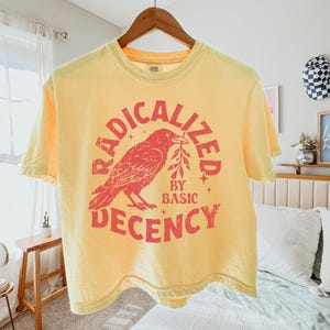 Könnte beinhalten: Ein blassgelbes Kurzarm-T-Shirt mit einem roten Grafikdruck eines Raben, der einen Zweig hält. Der Text RADICALIZED BY BASIC DECENCY ist in einer kreisförmigen roten Schrift um den Vogel herum auf einem Holzkleiderbügel abgebildet.