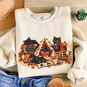 May include: A cream-colored crewneck sweatshirt featuring a graphic of three black cats reading books inside a small tent. The illustration includes pumpkins, a lantern, a ghost, and autumn leaves. The cats wear glasses and scarves.