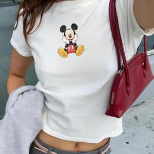 May include: White t-shirt with a Mickey Mouse graphic in black, red, and yellow. A red handbag with a zip closure hangs from the shoulder. The outfit includes grey trousers and a pink belt.