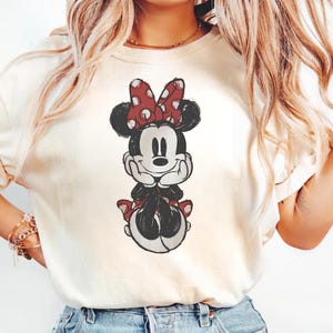 May include: Cream-coloured t-shirt with a distressed graphic of Minnie Mouse. Minnie is shown with a red polka-dot bow, black ears, and a red skirt. The design has a vintage, worn appearance.