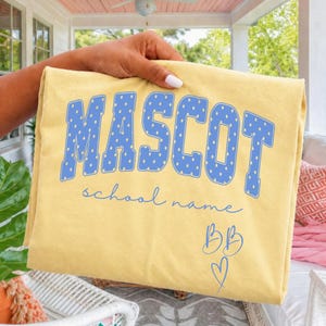 Pode incluir: Uma t-shirt amarelo-pálido com a palavra MASCOT em letras azuis grandes com um padrão de bolinhas brancas. Abaixo da palavra, o texto school name está impresso numa fonte cursiva azul, seguido pelas letras BB e um pequeno ícone de coração.