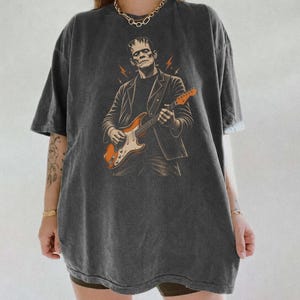 Puede incluir: Una camiseta gris carbón de corte oversize con un estampado gráfico de una figura que se asemeja al monstruo de Frankenstein tocando una guitarra eléctrica naranja. El diseño incluye pequeños rayos naranjas alrededor de la figura sobre la tela oscura.