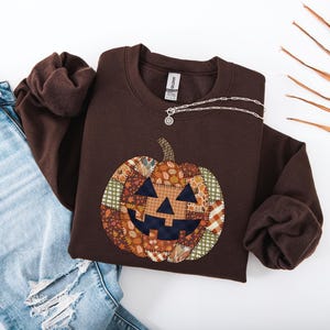 Op de afbeelding: Een donkerbruine sweater met ronde hals en een grote, centrale pompoenafbeelding. De pompoen is ontworpen met een patchworkpatroon van diverse herfstachtige stoffen, waaronder ruitjes, gingham en bladprints in tinten oranje, bruin en groen.