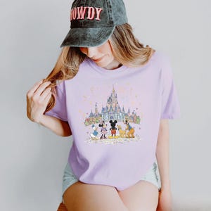 K&ouml;nnte beinhalten: Helllila T-Shirt mit einer farbenfrohen Illustration von Disney-Figuren vor einem Schloss. Das Design umfasst Micky Maus, Minnie Maus, Donald Duck, Goofy und Pluto. Das Shirt hat einen Rundhalsausschnitt.