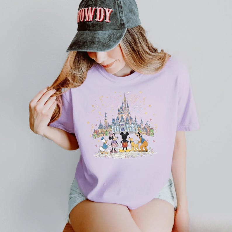 K&ouml;nnte beinhalten: Helllila T-Shirt mit einer farbenfrohen Illustration von Disney-Figuren vor einem Schloss. Das Design umfasst Micky Maus, Minnie Maus, Donald Duck, Goofy und Pluto. Das Shirt hat einen Rundhalsausschnitt.