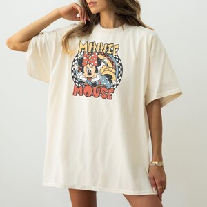 May include: Cream-colored oversized t-shirt featuring a graphic of Minnie Mouse. The design includes Minnie's face, a red bow, and the words "Minnie Mouse" in a retro font. The shirt has short sleeves and a relaxed fit.
