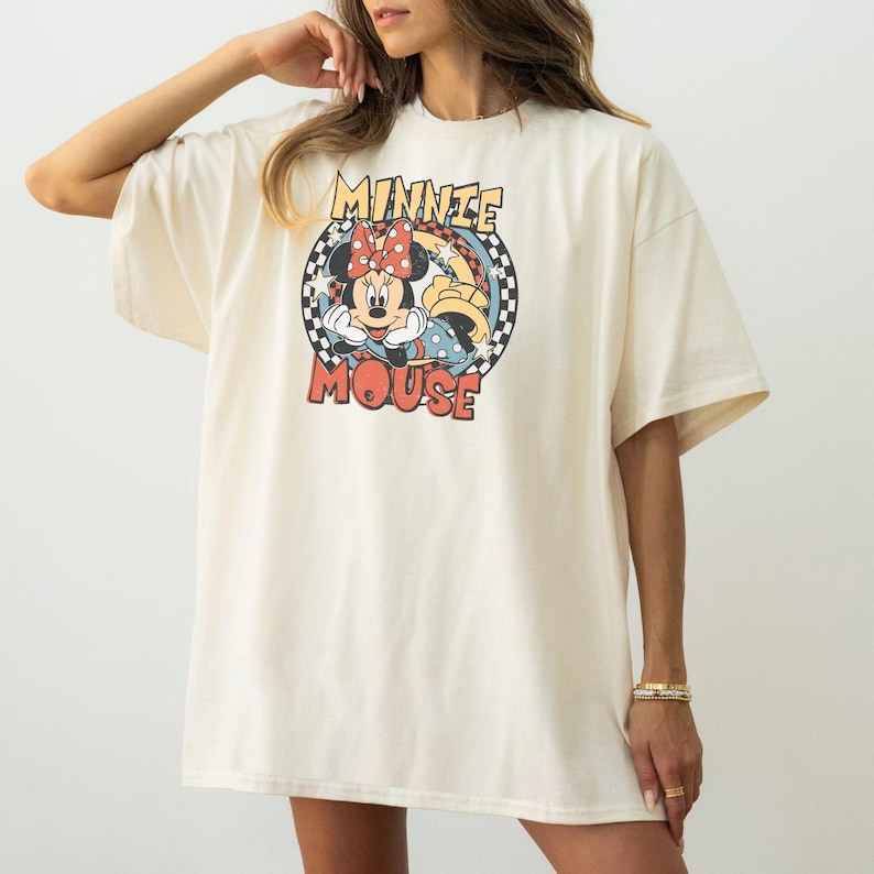 May include: Cream-colored oversized t-shirt featuring a graphic of Minnie Mouse. The design includes Minnie's face, a red bow, and the words "Minnie Mouse" in a retro font. The shirt has short sleeves and a relaxed fit.
