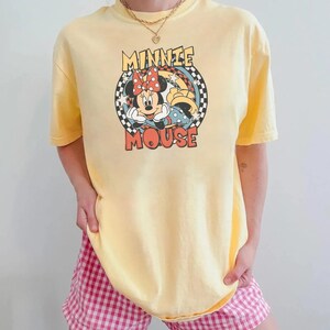 May include: Yellow t-shirt featuring a Minnie Mouse graphic. The graphic includes Minnie Mouse with a red bow, the words "MINNIE MOUSE", and a checkered background. The shirt has short sleeves and a crew neck.