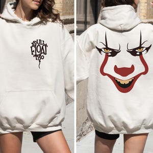 May include: An off-white hooded sweatshirt featuring a black graphic on the front chest that reads "YOU'LL FLOAT TOO" in a balloon shape. The back of the hoodie displays a large, detailed illustration of a clown face with red and black accents.