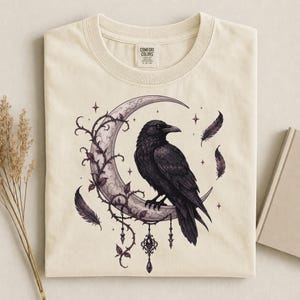 May include: A cream-colored t-shirt featuring a graphic print of a black raven perched on a crescent moon. The moon is entwined with thorny vines and dangling charms. Several dark feathers float around the moon, accented by small four-pointed stars.