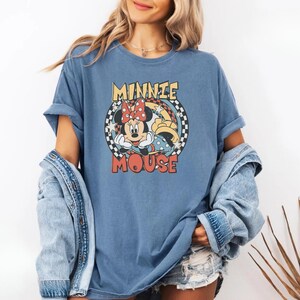 May include: Blue t-shirt featuring a graphic of Minnie Mouse with a red polka dot bow. The text "MINNIE MOUSE" is in a retro font. The shirt has rolled-up sleeves and a relaxed fit.