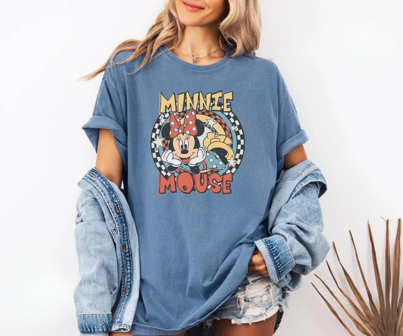 May include: Blue t-shirt featuring a graphic of Minnie Mouse with a red polka dot bow. The text "MINNIE MOUSE" is in a retro font. The shirt has rolled-up sleeves and a relaxed fit.