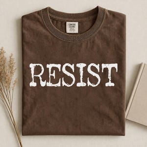 Op de afbeelding: Een donkerbruin t-shirt met korte mouwen en het woord RESIST in grote, witte, verweerde hoofdletters op de borst gedrukt. Het shirt heeft een ronde hals en een Comfort Colors merklabel dat zichtbaar is aan de binnenkant van de kraag.