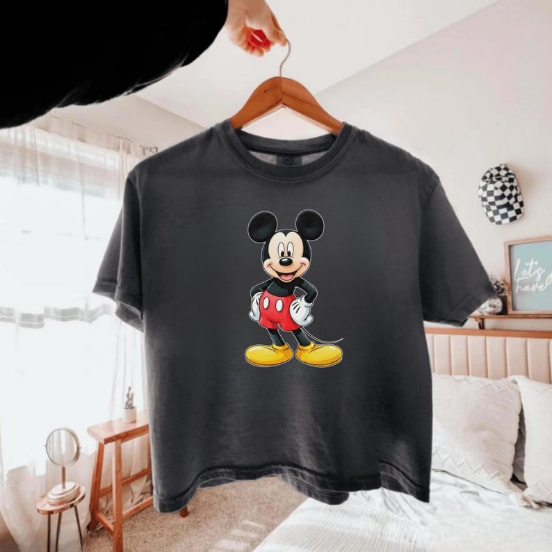 May include: A charcoal gray short-sleeve t-shirt featuring a centered graphic of Mickey Mouse. The character is depicted with his signature black ears, red shorts with two white buttons, white gloves, and large yellow shoes, standing on a plain background.