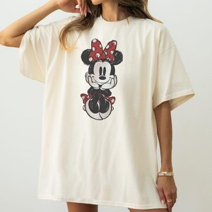 May include: Cream-coloured t-shirt featuring a distressed graphic of Minnie Mouse. The cartoon character is depicted with a red bow with white polka dots, a black face, and a red bow on her skirt. The shirt has short sleeves and a relaxed fit.