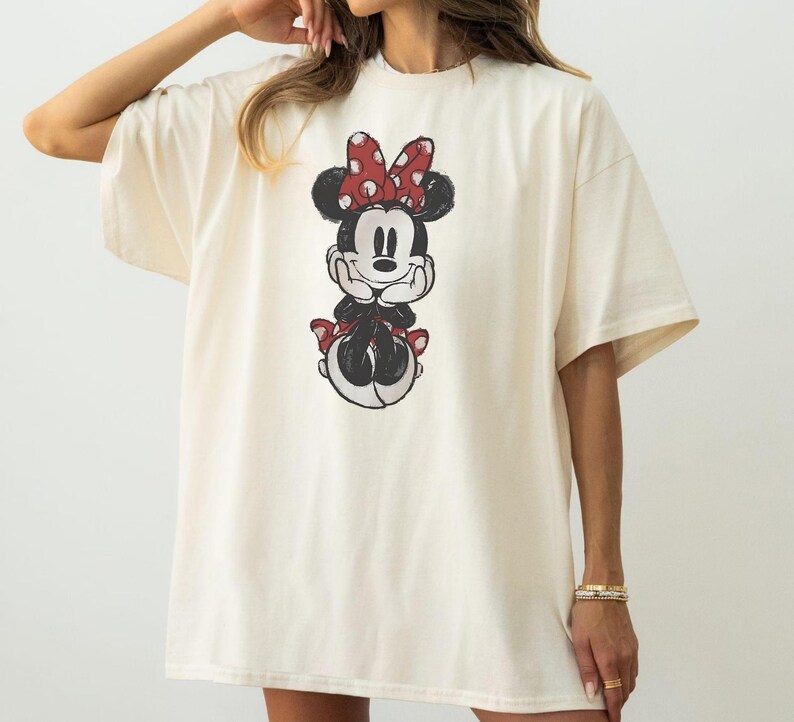 May include: Cream-coloured t-shirt featuring a distressed graphic of Minnie Mouse. The cartoon character is depicted with a red bow with white polka dots, a black face, and a red bow on her skirt. The shirt has short sleeves and a relaxed fit.