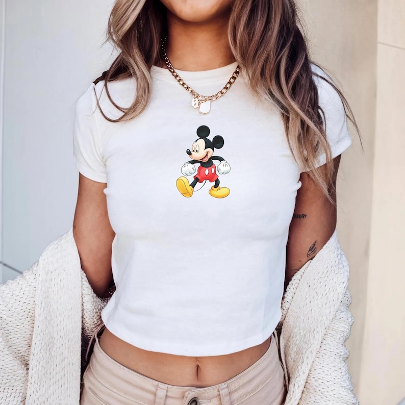 Puede incluir: Una camiseta blanca de manga corta con un gr&aacute;fico central de Mickey Mouse con pantalones cortos rojos y zapatos amarillos. La camiseta tiene cuello redondo y un corte ajustado. Se lleva un collar de cadena de tono dorado alrededor del cuello.