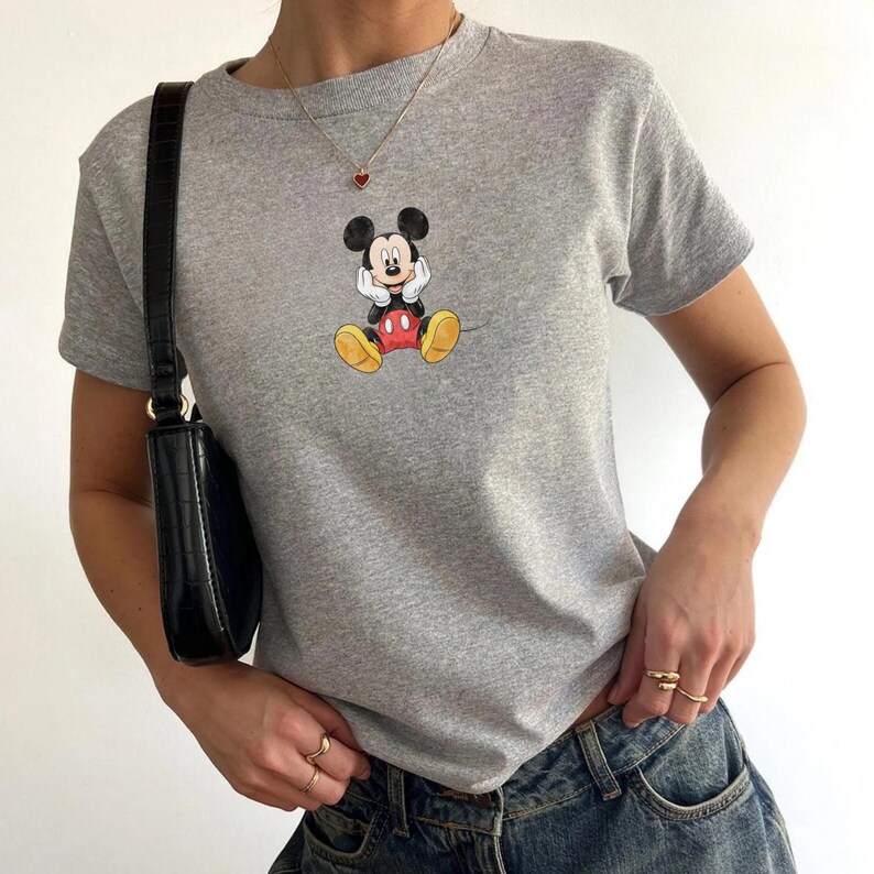 May include: A grey t-shirt with a Mickey Mouse graphic. The short-sleeved top has a crew neck and is worn with blue jeans. A black shoulder bag and gold rings complete the outfit.