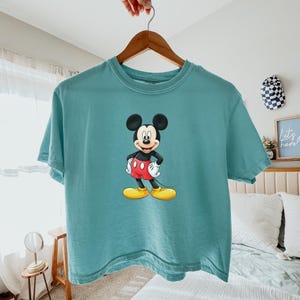 May include: A teal short-sleeve t-shirt featuring a centered graphic of Mickey Mouse. The character is depicted with his signature black ears, red shorts with two white buttons, white gloves, and large yellow shoes, standing in a classic pose.