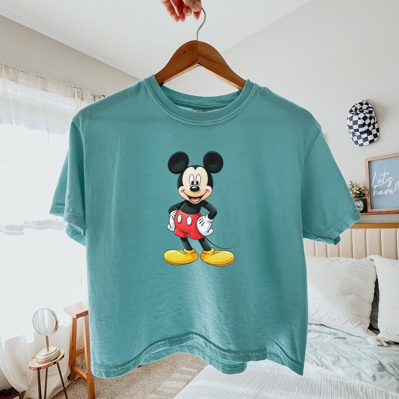 May include: A teal short-sleeve t-shirt featuring a centered graphic of Mickey Mouse. The character is depicted with his signature black ears, red shorts with two white buttons, white gloves, and large yellow shoes, standing in a classic pose.
