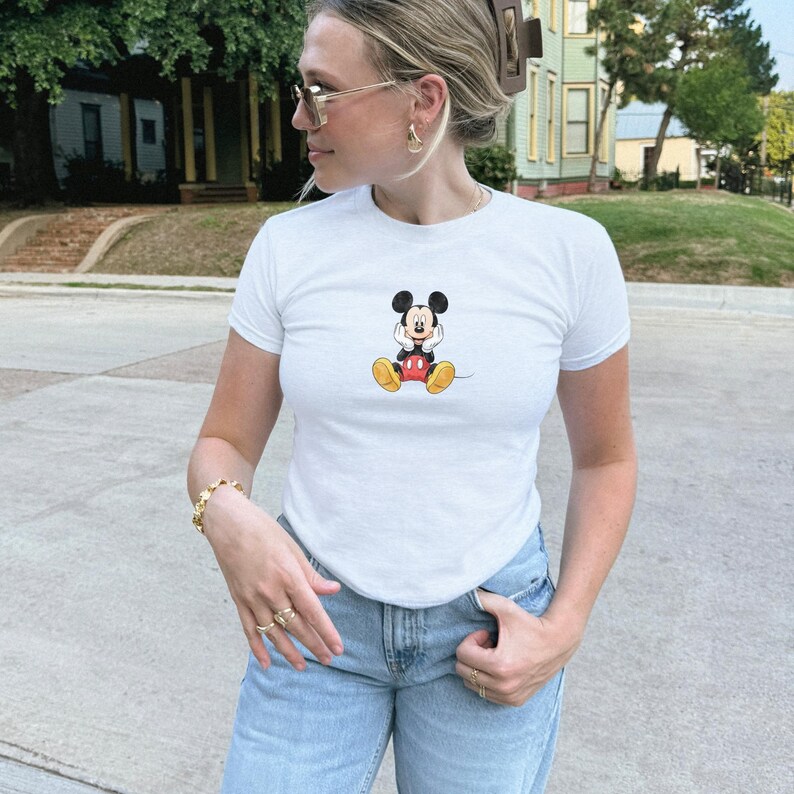 May include: White t-shirt with a small Mickey Mouse graphic. The cartoon character is shown in his classic pose, with red shorts, yellow shoes, and black gloves. The shirt is worn with light blue jeans.