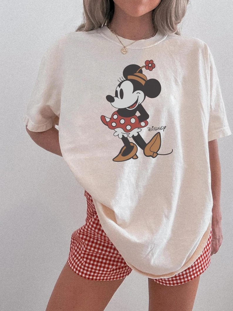 May include: Cream-colored t-shirt featuring a cartoon Minnie Mouse graphic in red, black, and tan. The word "Disney" is printed below the character. Paired with red and white checkered shorts.
