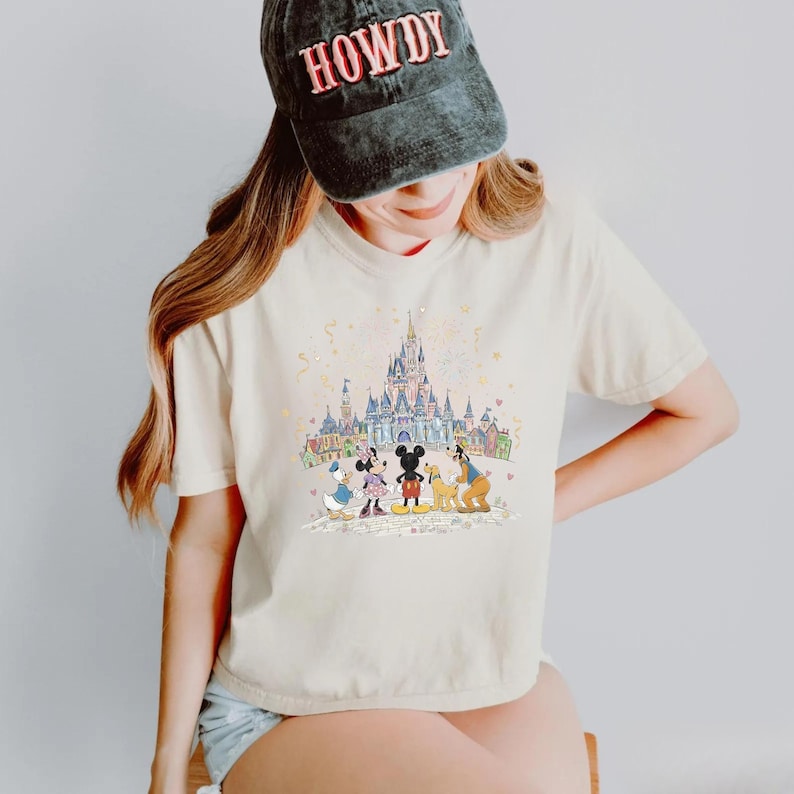 K&ouml;nnte beinhalten: Cremefarbenes T-Shirt mit einer farbenfrohen Illustration eines Schlosses und Zeichentrickfiguren. Eine schwarze Kappe mit dem Wort "HOWDY" wird getragen. Die Grafik zeigt Micky Maus, Minnie Maus, Donald Duck, Goofy und Pluto.