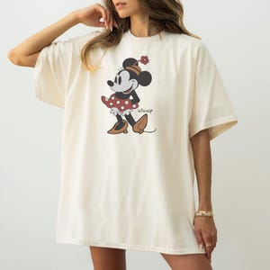 May include: Cream-colored t-shirt featuring a graphic of Minnie Mouse. The cartoon character is depicted in a red polka-dot dress, black shoes, and a flower on her head. The Disney logo is printed below the character.