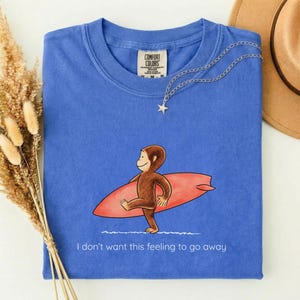 May include: A blue crew neck t-shirt with a Comfort Colors label at the inner neckline. The graphic features a brown monkey carrying a red surfboard, with the white text "I don't want this feeling to go away" printed below the illustration.
