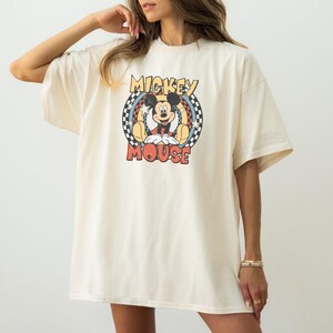 May include: Cream-colored t-shirt featuring a retro Mickey Mouse graphic. The design includes Mickey Mouse in a seated pose, surrounded by a circular design with the words "Mickey Mouse". The shirt has short sleeves and a relaxed fit.