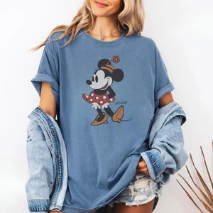 May include: A blue t-shirt featuring a Minnie Mouse graphic. The cartoon character is wearing a red polka dot skirt, a flower hat, and brown shoes. The word "Disney" is printed on the shirt.