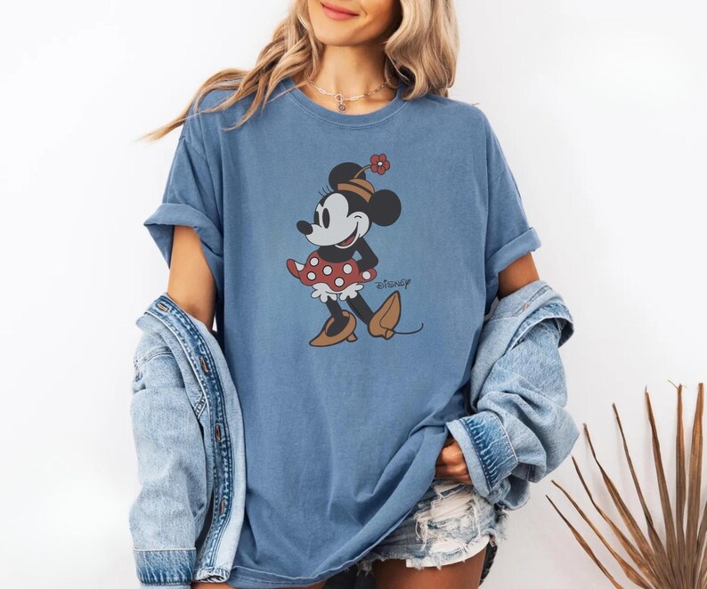 May include: A blue t-shirt featuring a Minnie Mouse graphic. The cartoon character is wearing a red polka dot skirt, a flower hat, and brown shoes. The word "Disney" is printed on the shirt.
